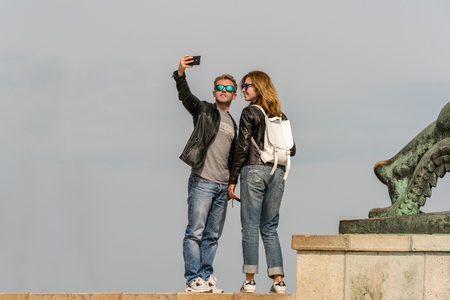Budapest, Hungary - September 26, 2017: Close up front view of a young caucasian tourist couple wearing sunglasses. Standing on a concrete  foundation at a viewpoint up high to take a panoramic selfie picture with their mobile phone of the city Budapest Hのeditorial素材