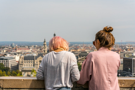 Budapest, Hungary - September 26, 2017: Close up back view of two young caucasian woman leaning over stone railing to relax and to look at the city view of Budapest Hungary in the background.のeditorial素材