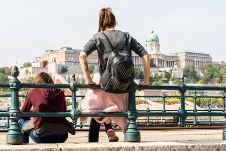 Budapest, Hungary - September 27, 2017: Low angle back view of two young caucasian women sitting and leaning against a steel railing by the Danube river in Budapest Hungary, resting and waiting for a tram. Buda Castle in the background.のeditorial素材