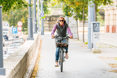 Budapest, Hungary - September 27, 2017: Selective focus front view of a caucasian woman wearing sunglasses riding a bicycle at an walk and cycle path in Budapest Hungary to avoid traffic. Autumn trees and traffic in the background.のeditorial素材