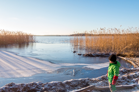 Young child in winter clothing looking at a beautiful cold sunny scenic tranquil winter landscape of ice, water and reed against a clear blue sky.の写真素材