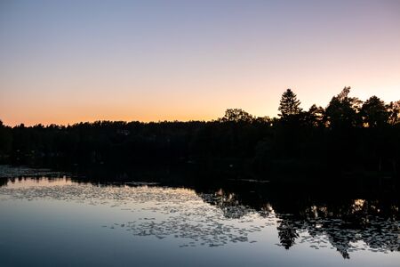 Beautiful late summer night sunset with calm water reflections and dark trees against clear sky.の写真素材