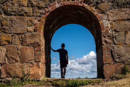 Summer view, silhouette of a young boy man leaning against an ancient old stone wall inside an arched gate looking at the blue sky. Varberg Fortress in Sweden.の写真素材