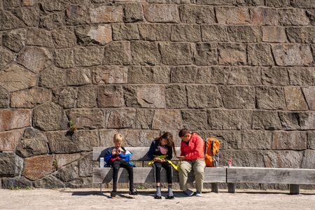 VARBERG, SWEDEN - JULY 16, 2019: Front view of one woman and two children sitting on a bench in front of a large stone wall at Varbergs Fortress looking at their smartphones in Varberg Sweden July 16, 2019.のeditorial素材