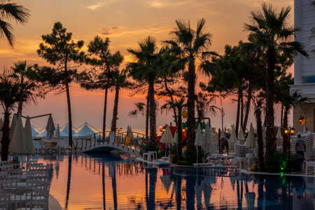 DURRES, ALBANIA - AUGUST 9, 2018: Beautiful sunset view of a pool area with palm trees, reflecting pool water and sun chairs with incidental people in the middle in Durres Albania August 9, 2016.のeditorial素材