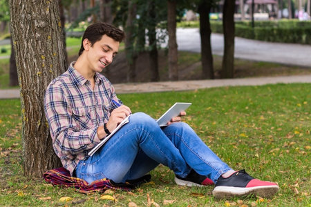 young men student sitting  in the park next to the tree with the tablet in his hands 