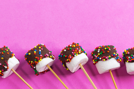 marshmallows on a sticks, laying on pink background. glazed with chocolate and colorful sprinkles.の写真素材