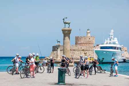 Rhodes, Greece - June 2021: Bicycle tour near Agios Nikolaos fortress at the entrance to Mandraki harbor and statue of deer on column in the foreground.のeditorial素材