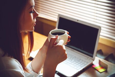 Portrait of beautiful young designer woman sitting at home office desk with cup of coffee and phoneの写真素材