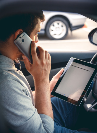 Young man driving a car with a tablet in hand, talking on mobile phone. Multitasking. Safe driving conceptの写真素材