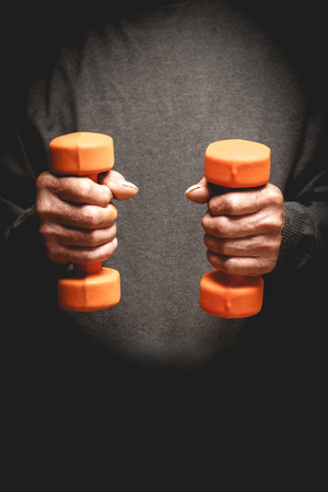 Orange dumbbells in the hands of an elderly man. Sports retired. Photo in low keyの写真素材