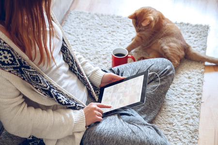 Woman in home cozy clothes sitting on the carpet near the sofa using a tablet with headphones,drinking coffee from a red cup. Next to it lies a thick red cat. Online education concept.の写真素材