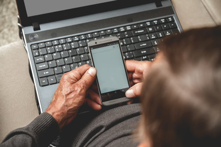 Senior sitting on a sofa with mobile phone in hand and laptop. Working in retirement concept