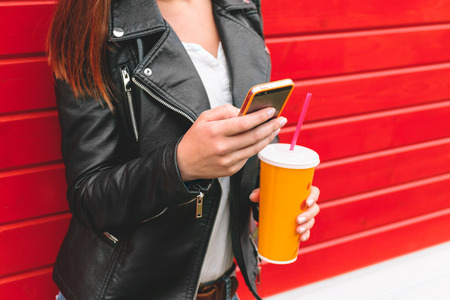 girl using mobile phone, writing SMS, with a drink in his hand on a red wooden wallの写真素材