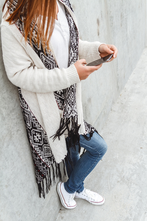 Portrait of a young beautiful girl with a phone hands  in the warmth of a knitted sweater in a concrete wallの写真素材