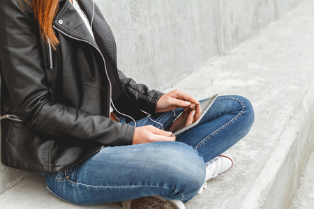 Girl with a tablet and headphones in hands sits on a concrete wall. Online education studentsの写真素材