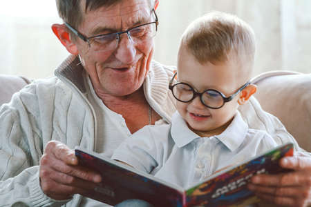 Elderly grandfather and little grandson read a book together and have fun sitting in a chairの写真素材