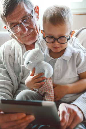 An elderly grandfather and his little grandson are using a tablet device together, smiling and...