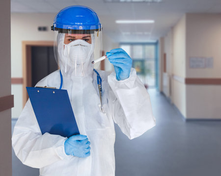 Female doctor or nurse in protective suit and face shield holding a positive blood test result for...