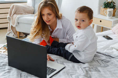 A young mother with her beloved son in the bedroom on the bed with a laptop. Work at home with a child conceptの写真素材
