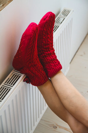 A young woman in warm, cozy knitted socks warms her legs by the radiatorの写真素材