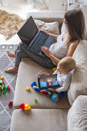 Young mom works at home sitting on couch with laptop while her little kid plays with tabletの写真素材