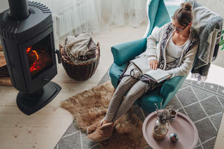 Young woman sitting in a cozy armchair with a warm blanket reading a bookの写真素材