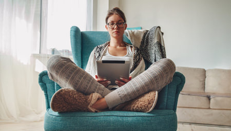Young woman sitting in cozy armchair, with warm blanket, using tablet