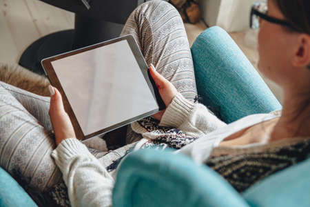 Young woman sitting in cozy armchair, with warm blanket, using tabletの写真素材