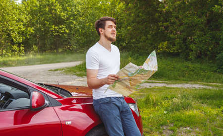The man with the map in hand standing next to a car in the forest. Get lost. Car travel, hitchhiking conceptの写真素材