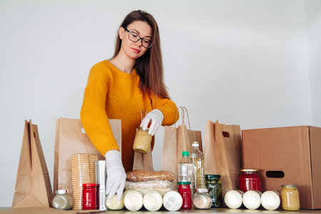 Young woman volunteer wearing medical gloves packing food donation in paper bag