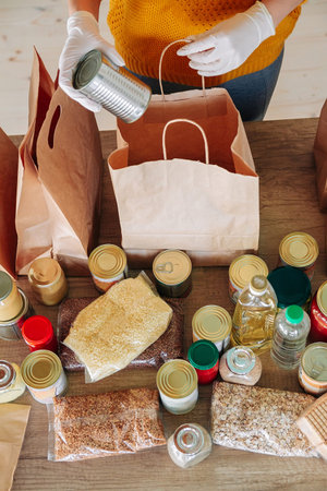 Young woman volunteer wearing medical gloves packing food donation in paper bag