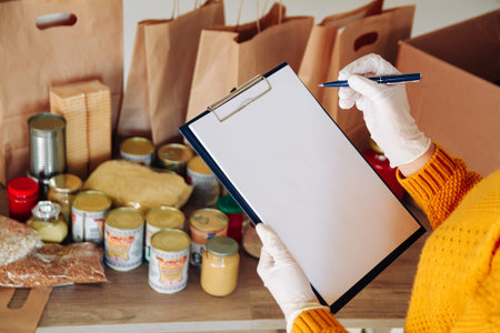 Young woman volunteer in medical gloves writes down and packs food for donation in paper bag