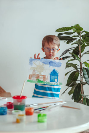 Little child boy paints with watercolors while sitting at the table, at homeの写真素材