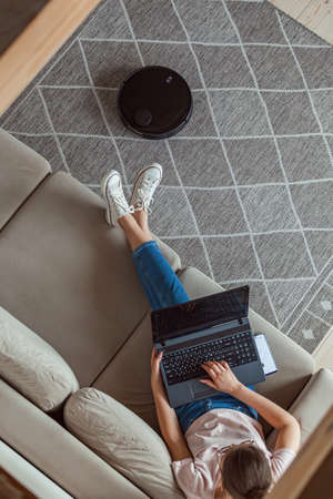 Robot vacuum cleaner cleaning carpet, woman using laptop sitting on the sofa at homeの写真素材