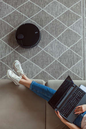 Robot vacuum cleaner cleaning carpet, woman using laptop sitting on the sofa at homeの写真素材