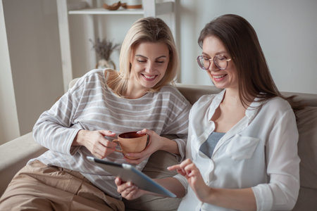 Two young women friends using tablet together, sitting on the couch, smiling and fun