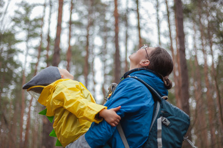 Happy family Mom and child walk in the forest after rain in raincoats together, hug and look at the skyの写真素材