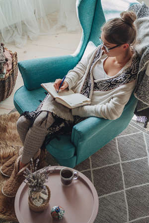 Young woman write in notebook sitting in armchair by fireplaceの写真素材