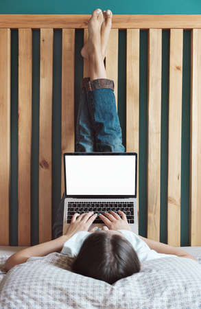 Young woman using laptop blank screen to work, lying on the bed in the bedroom, legs up. Flexible hours and remote work.の写真素材
