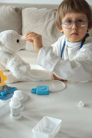 Little doctor in medical coat with stethoscope play eye treatment to teddy bear toyの写真素材