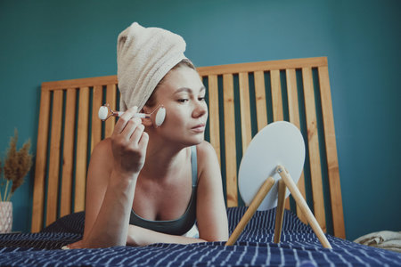 Young woman using jade facial roller for face massage lying on bed in bedroom looking in the mirror