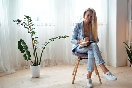 Young woman making notes in notebook sitting on a chair at home,look at cameraの写真素材