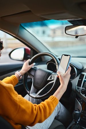 Hands of unrecognizable woman driver using white screen mockup mobile phone while driving