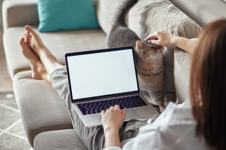 Mockup white screen laptop woman using computer and pet cat lying on sofa at home, back view, focus on screenの写真素材