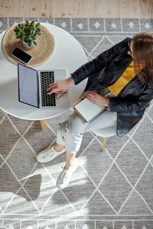 Young woman using laptop and making notes in notebook sitting at workplace at home, remote work top viewの写真素材