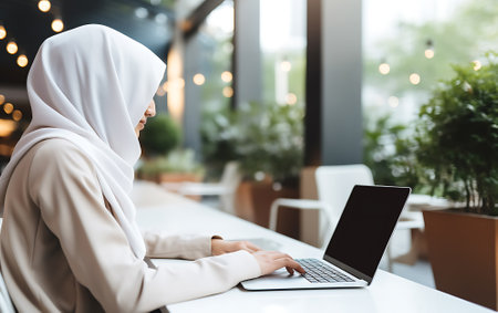 Young arab woman in hijab using blank screen laptop in cafe, remote work concept.の素材