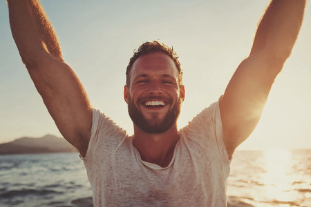 A joyful man with a beard raising his arms in celebration, standing on a beach during a beautiful sunrise.の素材