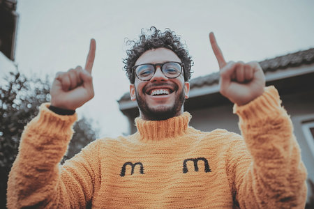 Joyful portrait of a young man with curly hair and glasses, wearing a bright yellow sweater. He raises his arms in a celebratory pose, radiating happiness and confidence in an open outdoor setting.の素材
