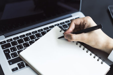 Closeup of man hand writing in diary on a table with laptop. Focused on a pen